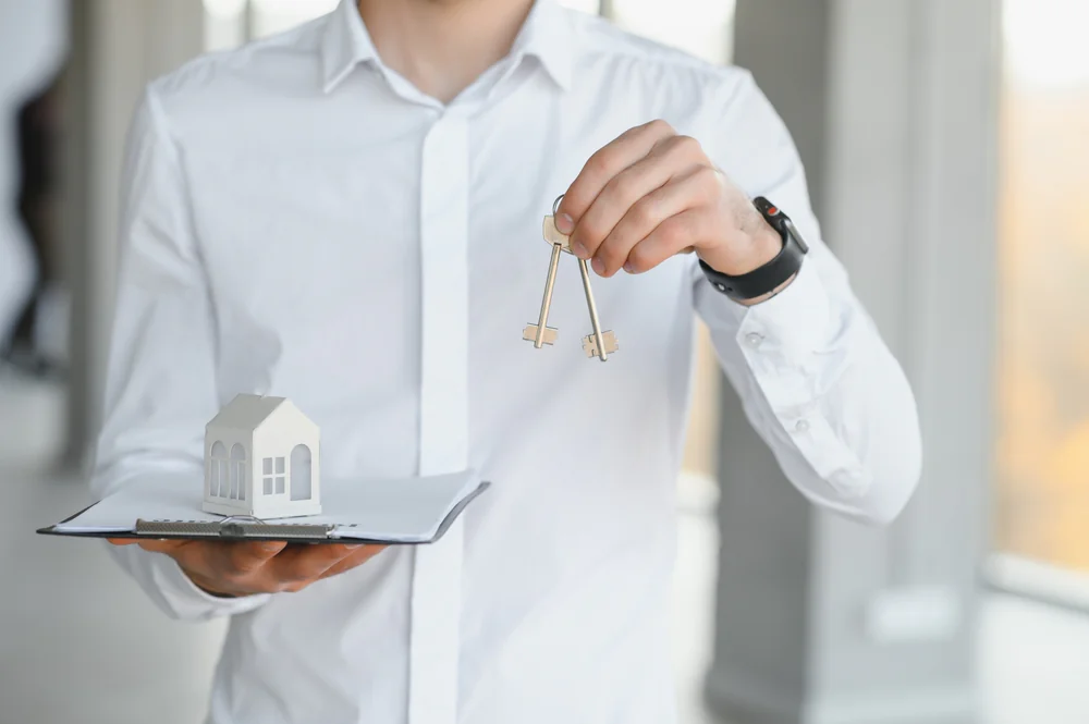 A person holds the keys to a property over a clipboard with a small house model
